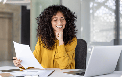 Image of a woman with a piece of paper in her hand smiling at laptop Image of a woman with a piece of paper in her hand smiling at laptop